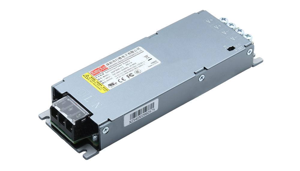 Gray metal DC power supply module with a screw-terminal block on one end and side mounting ears; labeled V+ and V- terminals and safety stickers