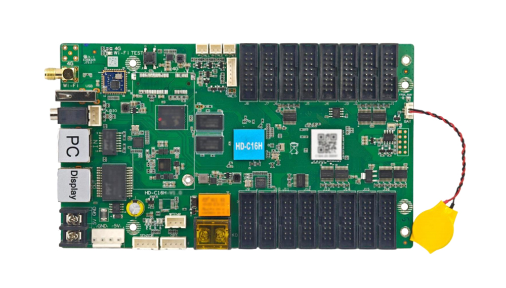 Green development board with numerous edge connectors and two labeled ports (PC and Display), plus a yellow battery pack on a red/black wire on the right side.